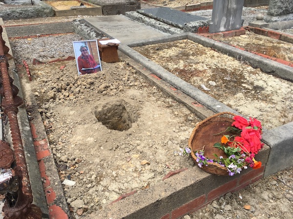 a grave plot with a small hole dug in it, with a basket of flowers and a photo of an elderly woman