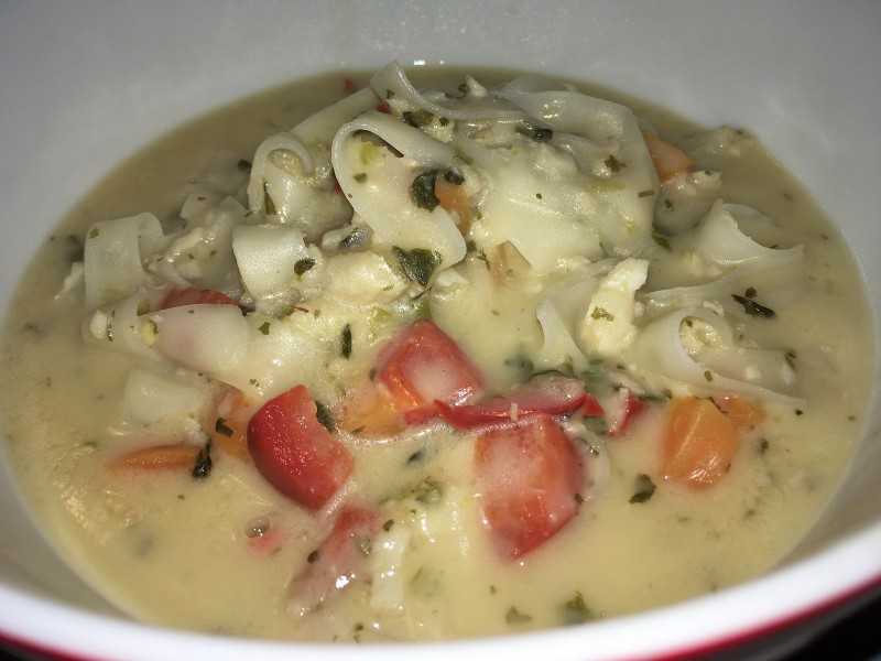Close-up of a bowl of light-coloured soup, with some rice noodles, red capsicum and carrot identifiable