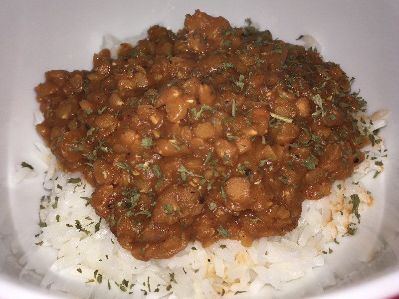 A close-up picture of a reddish lentil-based dish, atop a bowl of rice.