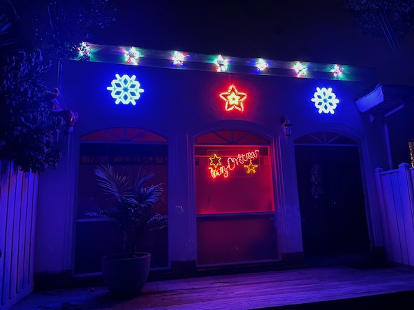 two older-style houses side-by-side, the left one with a moderate row of white lights on the eaves, the right one completely decked out in blaring lights