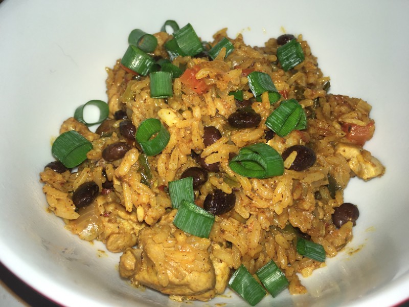a close-up of a dish with chicken, rice, beans, capsicum, spring onion all mixed together, tinted orange by spices, in a white-coloured bowl