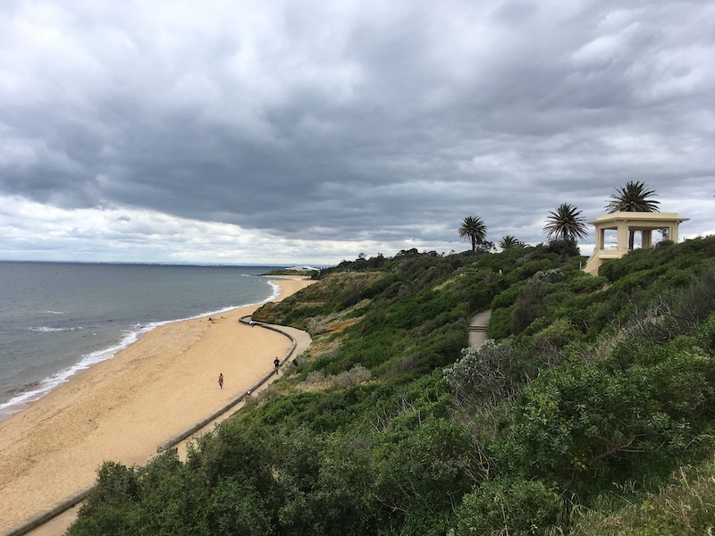 a view from a coastal cliff; greenery and a white structure to the right, sand and ocean to the left, and cloudy skies above