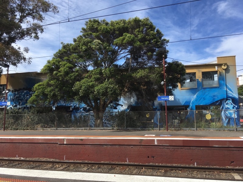 in the foreground, a train platform, behind which is a large tree, and behind that is a two-storey building with a large blue mural
