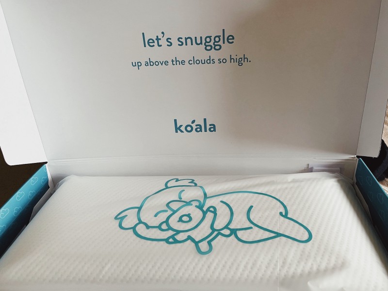 a photo of a cardboard box, opened. on the underside of the lid it says 'let's snuggle up above the clouds so high. koala'. in the box itself, there's a white pillow wrapped in plastic; the plastic has an illustration of a koala snuggling a teddy bear