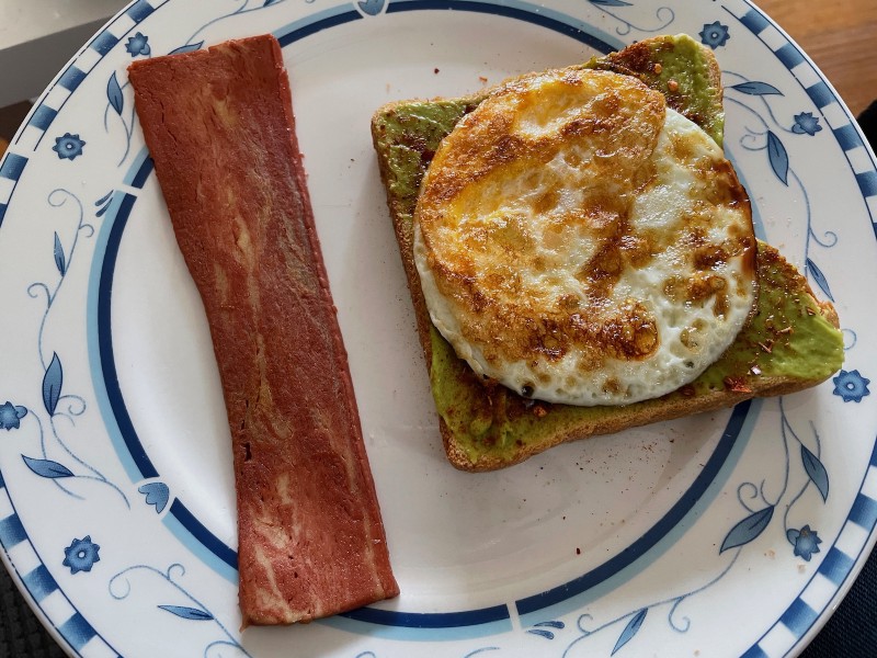 a plate with a fried egg on avocado toast and a slice of imitation bacon on it