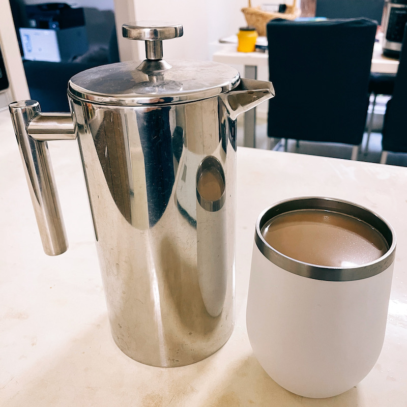 a photo of a metal coffee plunger and a white tumbler of white coffee, on a white bench