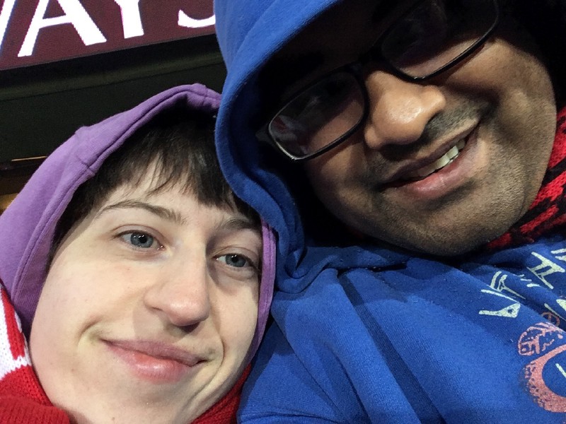 a white woman in a purple hooded coat and a Sydney Swans scarf, sitting close to an Indian man in a blue hoodie and (just visible) Essendon Bombers scarf, both smiling at the camera