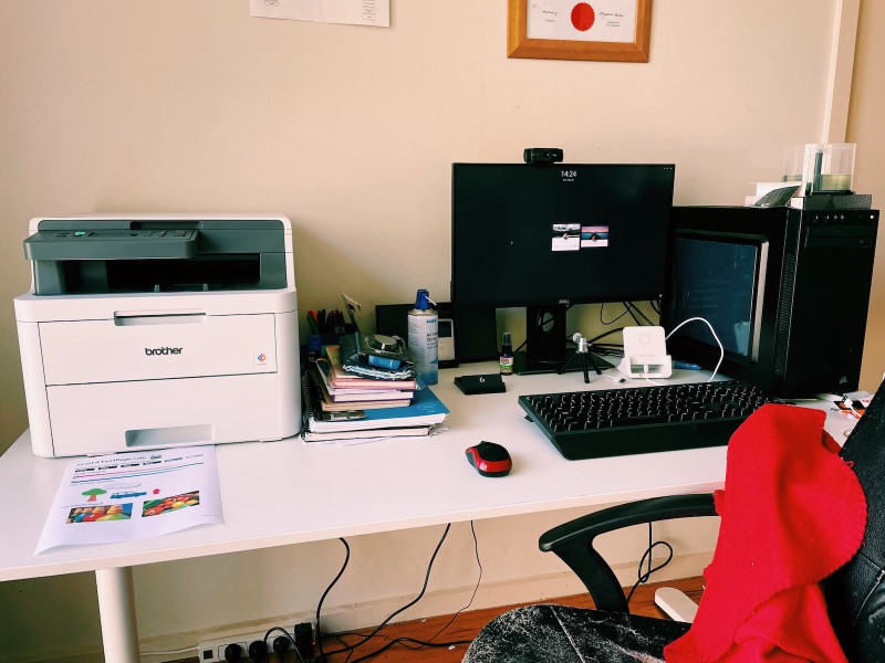 a computer desk, with a printer to the left, desktop computer to the right, and a stack of papers and notebooks in between