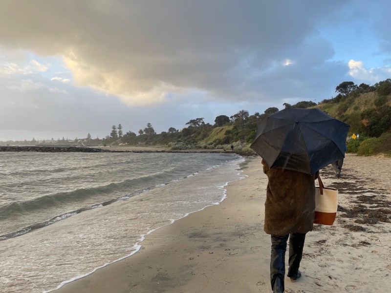 photo of a young woman from behind, holding an umbrella, on a beach in the sun and rain simultaneously