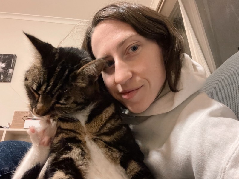 a young woman in a white hoodie takes a selfie with a tabby cat, whose hind leg is outstretched and toes extended so she can gnaw at the gap