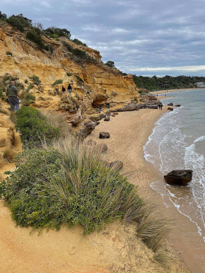 a reddish cliff seen side-on with yellow sand and ocean nearby
