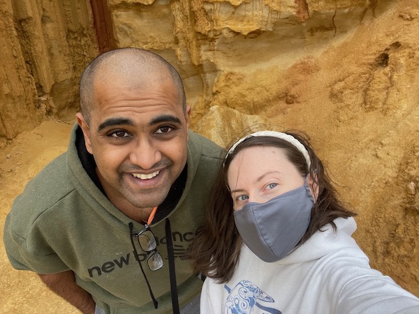 selfie of a masked white woman and unmasked Indian man, reddish cliff-face behind them