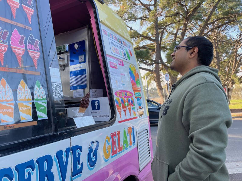 an Indian guy in a green hoodie stands at the side of an ice cream truck, grinning, as native Australian trees tower over him in the background