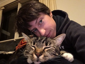 a selfie of a woman in a hoodie kissing the top of a tabby cat's head